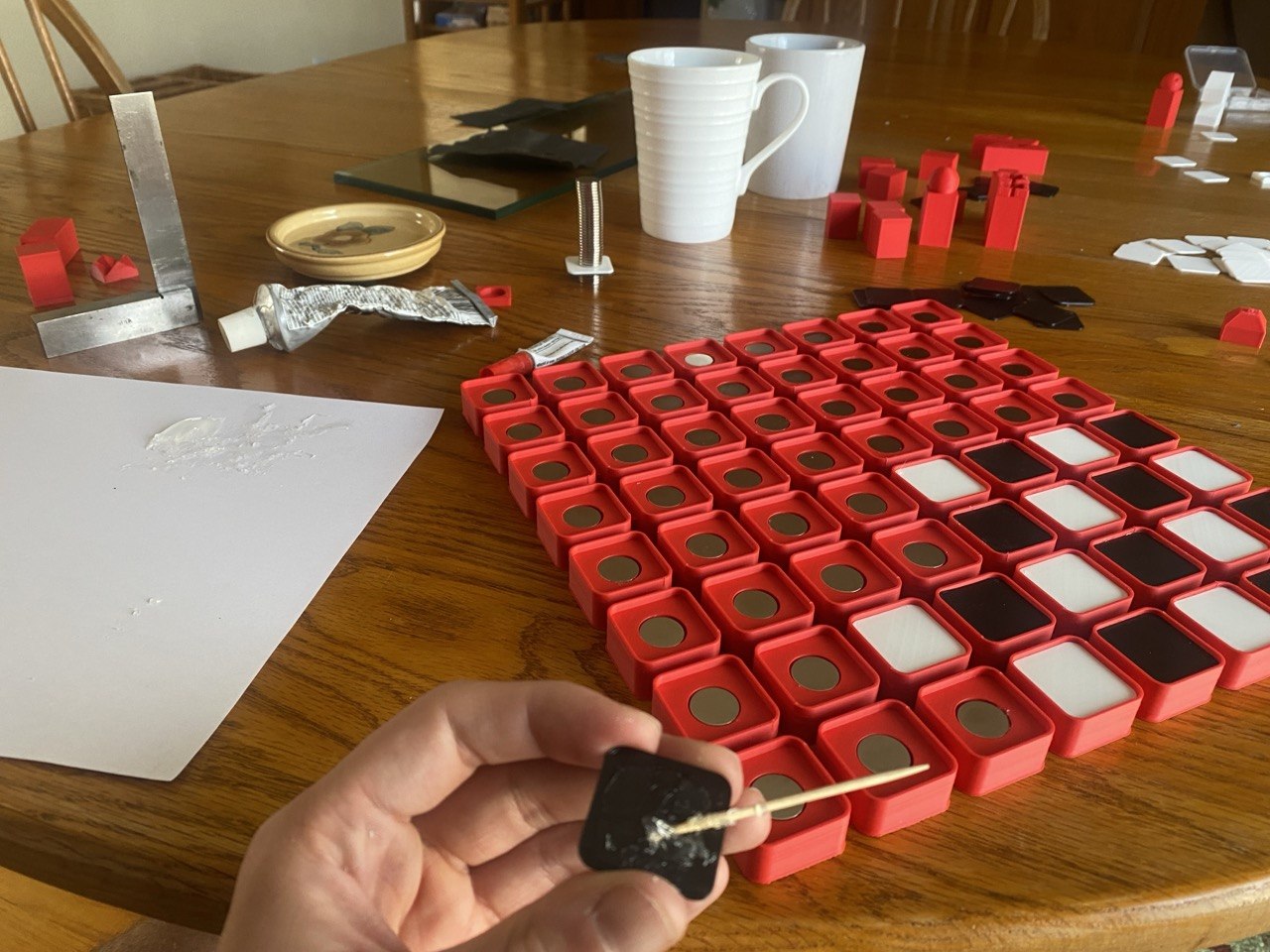 Assembly underway on the table: the red board with a magnet seated in each square's pocket, a tube of E6000, a machinist's square, and a black square top being glued on with a toothpick.