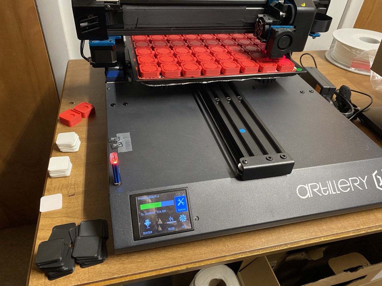 The Artillery Sidewinder mid-print, its bed packed with a grid of red board-base squares, with stacks of finished white, black, and red square tops resting beside it.